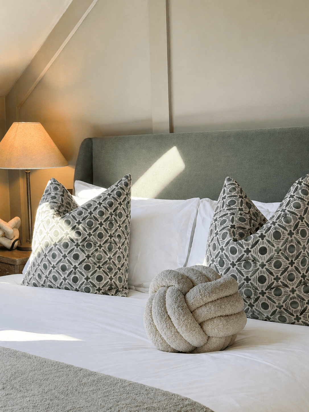 A styled bedroom scene showing a bed with white linens, two gray-and-white patterned accent pillows, a beige knotted decorative pillow, a gray upholstered headboard, and a bedside lamp with a warm lit shade.