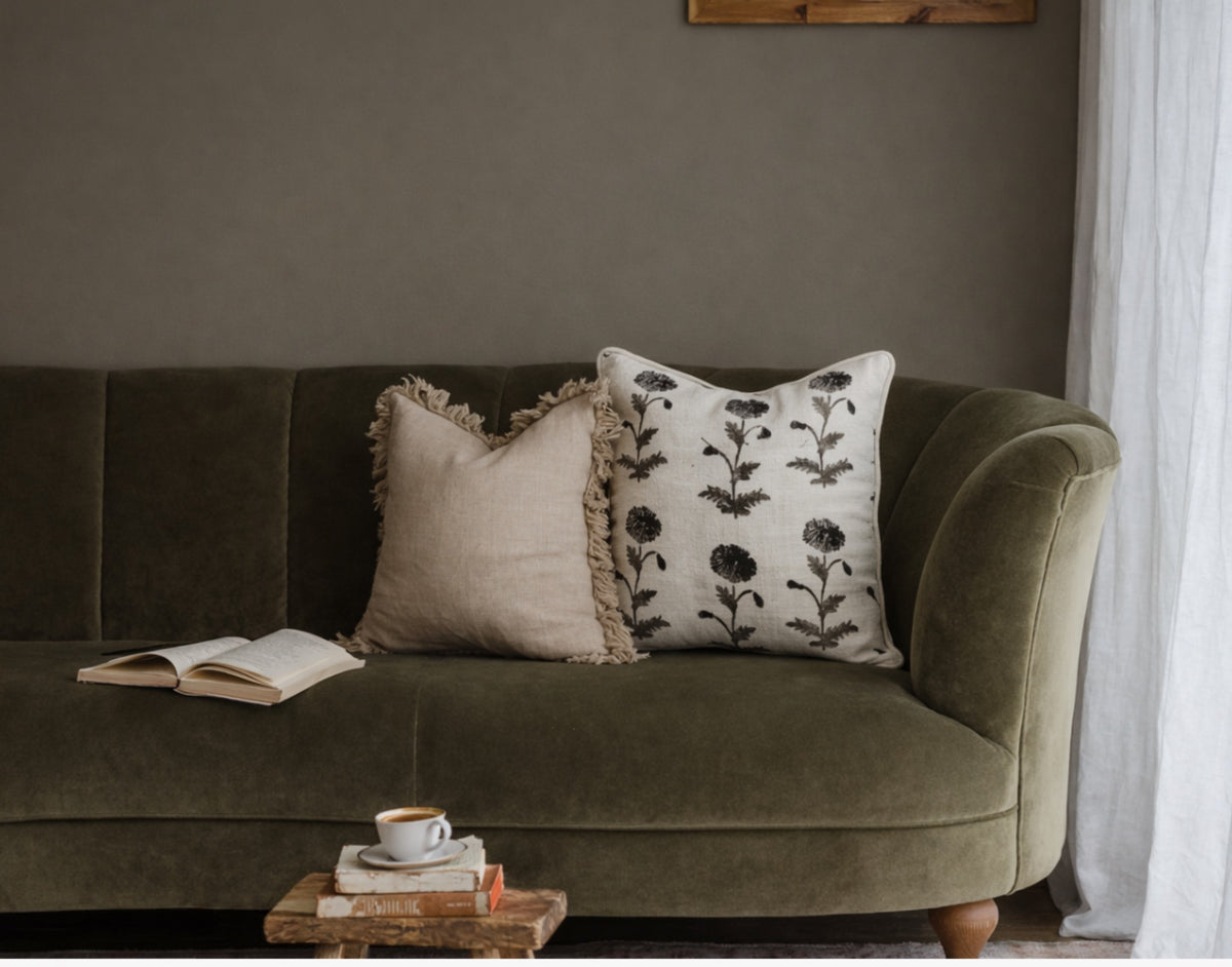 An olive green velvet sofa in a cozy living room setting, styled with two decorative throw pillows, an open book, and a small stack of books with a cup on a wooden tray in the foreground.