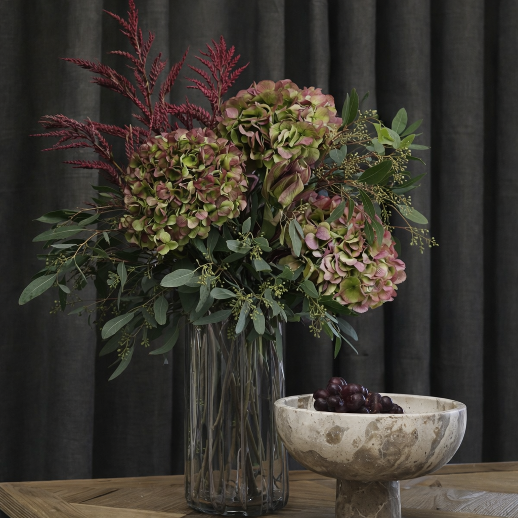 A floral arrangement of pink and green hydrangeas with burgundy feathery stems in a tall ribbed glass vase, placed on a wooden table beside a marble pedestal bowl containing dark grapes, set against a dark curtain background.